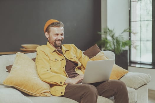 Smiling young man enjoys comfort of home while working on laptop. Laptop on knees, exemplifies modern lifestyle of remote work and telecommuting, showcasing perfect balance between work and relaxation. High quality photo