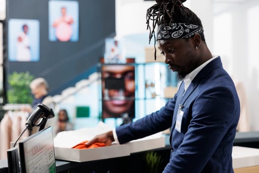 African american employee standing at cash register, preparing customer packages. Showroom worker putting fashionable clothes in carton boxes, working in shopping centre. Fashion concept