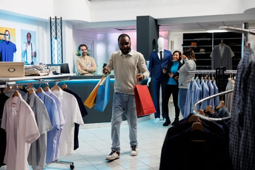 Happy shopaholic carrying paper bags filled with apparel in clothing store. Excited african american man holding packages while shopping and making purchase in mall boutique