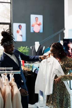 Boutique worker helping client with formal wear, looking at white shirt discussing material in clothing store. African american customer buying maternity clothes and trendy merchandise