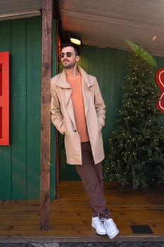 Man wearing sunglasses posing outdoors near Christmas tree. Male wearing stylish trench coat standing hands in pocket pose near building. Gentleman in relaxed pose during vacation