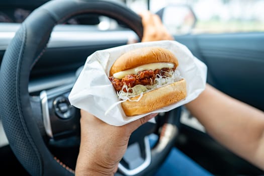 Asian lady holding hamburger to eat in car, dangerous and risk an accident.