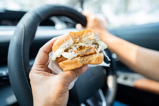 Asian lady holding hamburger to eat in car, dangerous and risk an accident.