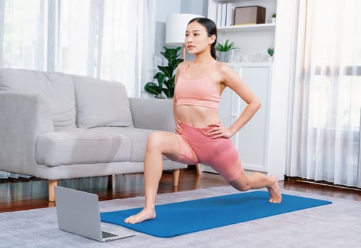 Asian woman in sportswear doing yoga exercise on fitness mat as her home workout training routine. Healthy body care lifestyle woman watching online yoga video on laptop. Vigorous