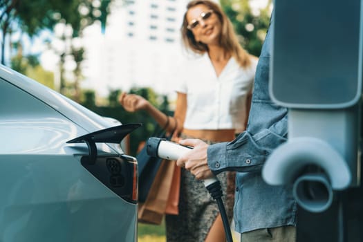 Young couple travel with EV electric car charging in green sustainable city outdoor garden in summer shows urban sustainability lifestyle by green clean rechargeable energy of electric vehicle innards