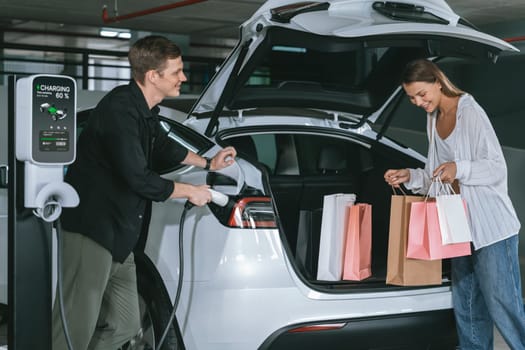 Young couple travel with EV electric car to shopping center parking lot charging in downtown city showing urban sustainability lifestyle by green clean rechargeable energy of electric vehicle innards