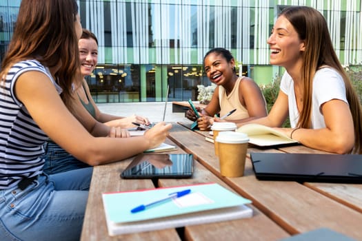 Group of multiracial female university students laughing, enjoying time together outdoor college campus studying and doing homework using laptops. Education and friendship concept.