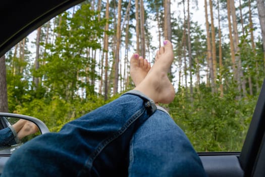 Female legs in blue jeans out of car window. Concept of comfortable local travel vacation holiday. Reduce carbon footprint. Woman resting in road trip refresh and recharge