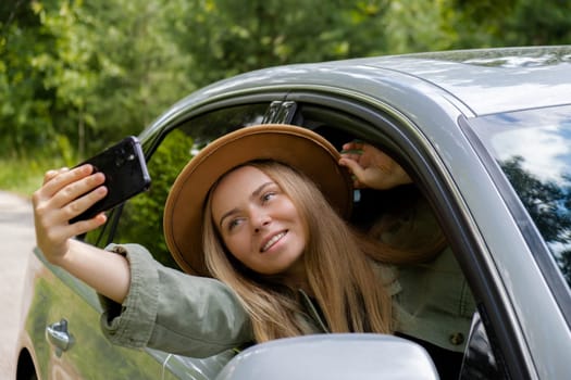 Exited smiling woman making video call with mobile phone from car window. Local solo travel on weekends concept. Young traveler explore freedom outdoors in forest taking selfie photo. Unity with nature lifestyle, rest recharge relaxation