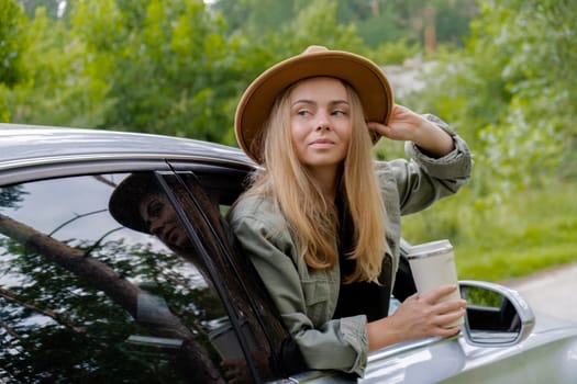 Blonde woman sticking head out of windshield car and drink coffee or tea from reusable mug. Young tourist explore local travel making candid real moments. Refuse reuse recycle zero waste concept. Responsible traveling reduce carbon footprint sustainable lifestyle