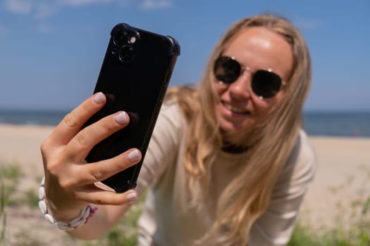 Young woman in sunglasses with shell bracelets holding smartphone. Influencer Using mobile phone for video call selfie waving on the beach seacoast. Holiday vacation advertisement concept. Social media