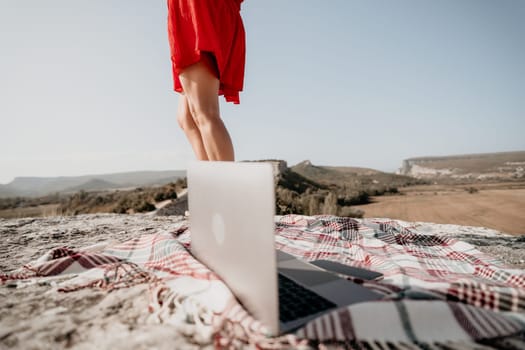 A laptop sits on a plaid blanket spread out on a rocky cliff with a mountain view in the background, with a woman in a red dress standing further back.