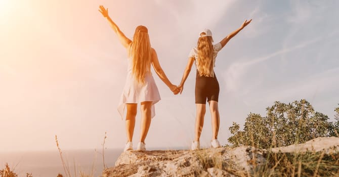 Two women, a mother and her daughter, stand on a cliff overlooking the ocean, hands raised in the air, as if in celebration.