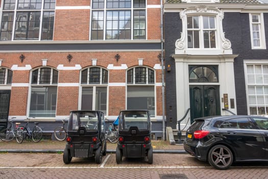 Netherlands. Summer day in Amsterdam. Two electric mini cars, a regular car and some bicycles are parked near an old building