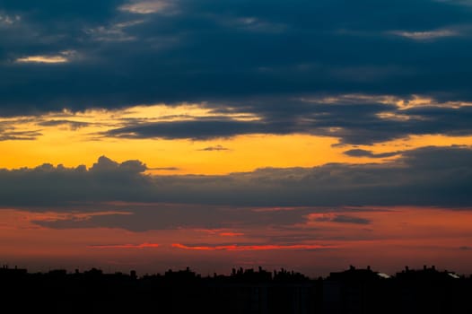 Cloudy sky above the city rooftops. The sun, just hidden behind the silhouettes of the buildings, beautifully colors the sky