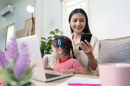 A mother multitasking with her daughter, working on a laptop and using a smartphone in a cozy home office environment.