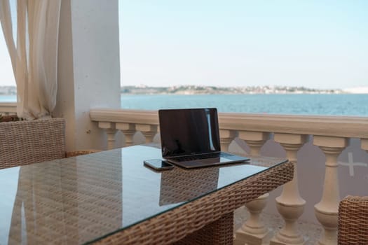 A laptop and a cell phone are on a table by a beach. The scene is peaceful and relaxing, with the ocean in the background. The table is made of clear plastic