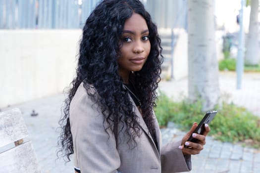 Beautiful black woman with curly hair using her smartphone to chat with friends looking at camera. connected concept