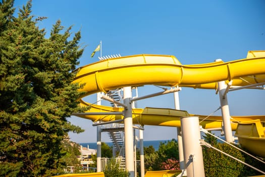 A bright yellow water slide curves through the air, supported by white poles against a blue sky. Lush green trees surround the base of the slide. family vacation concept