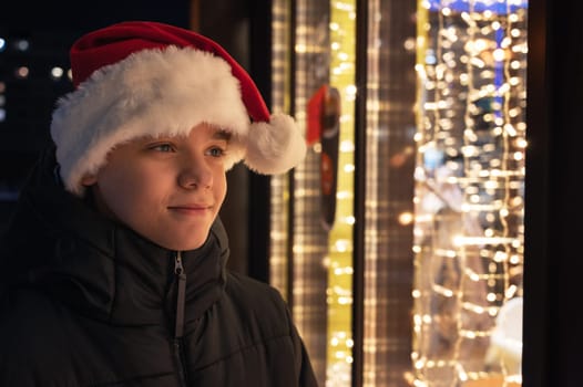 Boy in Santas hat looking and dreaming in illuminated shop window. Xmas presents holidays, or shopping on New Year or Christmas concept