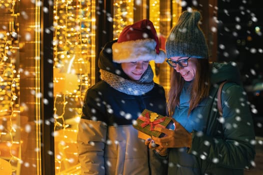 Pretty woman and her son in santas hat opening gift box on illuminated shop window background. New Year or Christmas concept