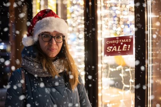 Pretty Woman in Santas hat looking and dreaming in illuminated shop window. Christmas holidays sales concept
