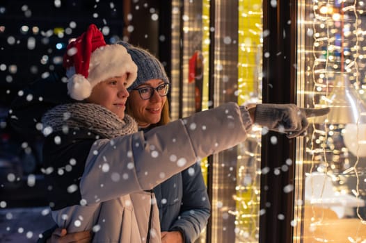 Pretty Woman and her son in santas hat looking and dreaming in illuminated shop window. Xmas presents holidays, or shopping on New Year or Christmas concept