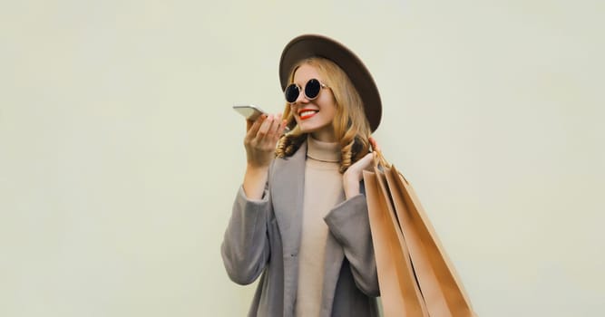 Beautiful happy young woman calling on smartphone with shopping bag, pretty girl smiles wearing hat, glasses standing on brown background