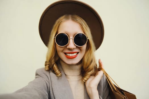 Autumn portrait of beautiful happy smiling young woman taking selfie with smartphone holding shopping bags in glasses, round hat
