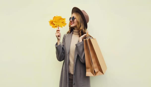 Autumn portrait of beautiful happy young woman with shopping bag, stylish girl holding yellow maple leaves in round hat, coat on brown studio background