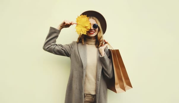 Autumn portrait of beautiful happy young woman with shopping bag, stylish girl holding yellow maple leaves in round hat, coat on brown studio background