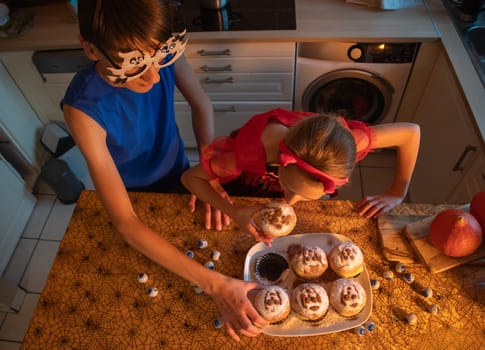 girl and brother trying buns with ghost images, they are dressed in Halloween costumes, enjoying holiday treats in decorated dark kitchen, warm cozy atmosphere, holiday mood,