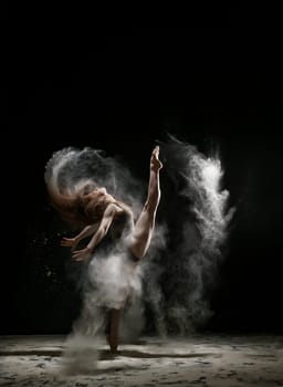 Female sexy gymnast in bodysuit shot in a jump on black background in a cloud of white dust all over body