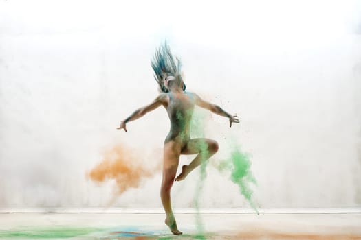 Female sexy gymnast in gray bodysuit shot in a jump on white background in a cloud of orange and green dust all over body