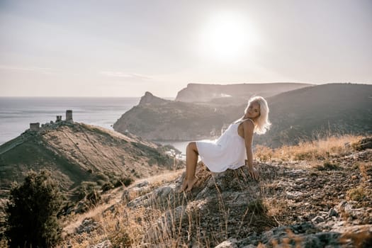 A woman is sitting on a hillside overlooking the ocean. She is wearing a white dress and has blonde hair. The scene is serene and peaceful, with the ocean in the background