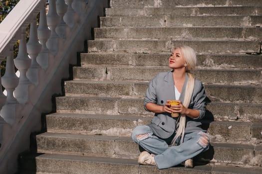 A woman sits on a set of stairs, holding a cup of coffee. She is enjoying her coffee and taking in the view from the top of the stairs