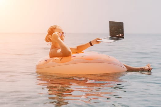 Woman freelancer works on laptop swimming in sea on pink inflatable ring. Pretty lady typing on computer while floating in the sea on inflatable donut at sunset. Freelance, remote work on vacation