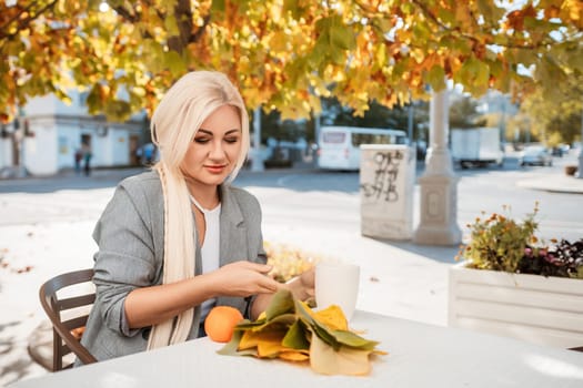 A blonde woman sitting at a table with a cup of coffee and a plate of food. The table is surrounded by potted plants and there are oranges on the table