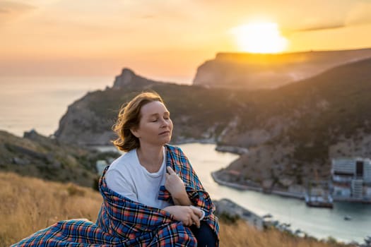 A woman is sitting on a hill with a blanket wrapped around her. She is looking out at the ocean