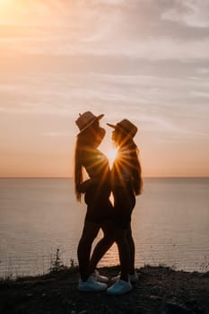 Two young women in hats stand side by side, silhouetted against a golden sunset over the ocean.