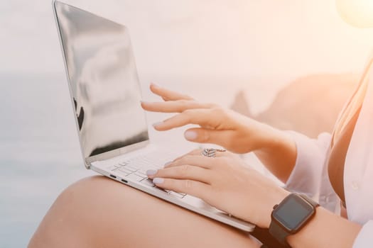 Digital nomad, woman in the hat, a business woman with a laptop sits on the rocks by the sea during sunset, makes a business transaction online from a distance. Freelance, remote work on vacation.