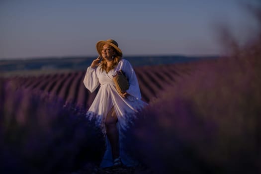 Woman poses in lavender field at sunset. Happy woman in white dress holds lavender bouquet. Aromatherapy concept, lavender oil, photo session in lavender.