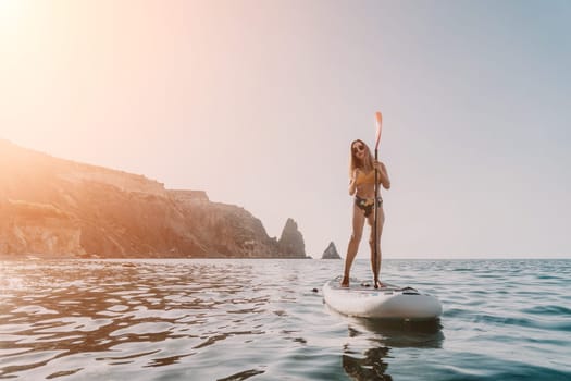 A woman in a bikini paddleboards on a calm sea with rocky cliffs in the distance.
