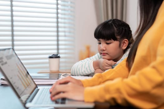Mother works on her laptop as her daughter studies close by, emphasizing the bond and balance between work and homeschooling.