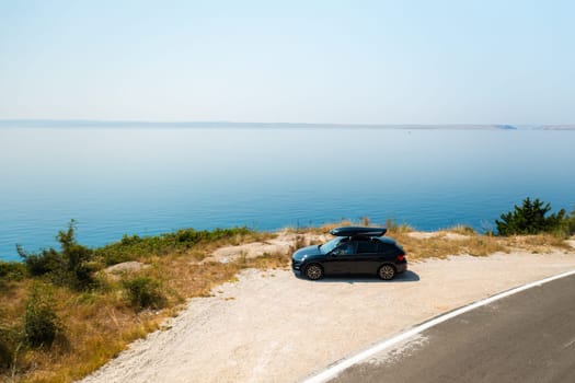 Sport car with a roof box, parked near a sharp turn on a coastal cliff road, with the azure sea below