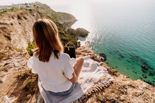 A woman is sitting on a rock overlooking the ocean with a laptop in front of her. She is enjoying the view and the peacefulness of the location