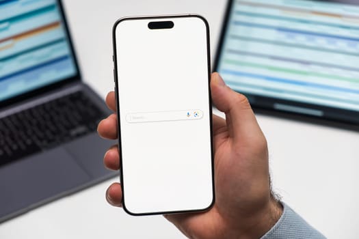 PRAGUE, CZECHIA - JULY, 2024: Google Search logo on the screen of smartphone in mans hand and laptop on the background