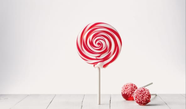 A large lollipop with red and white swirls stands upright on a white table. A pink triangle cast by a light source is behind it. Two smaller red lollipops with white sprinkles are laying on the table to the right of the larger lollipop. The background is a white wall.