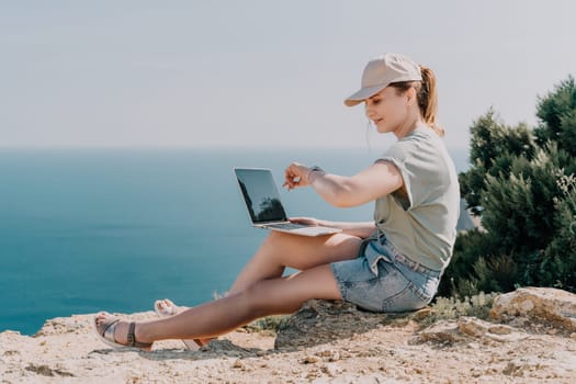 A young woman in a hat and casual clothes sits on a cliffside with a laptop, overlooking the ocean.