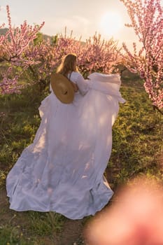 Woman blooming peach orchard. Against the backdrop of a picturesque peach orchard, a woman in a long white dress and hat enjoys a peaceful walk in the park, surrounded by the beauty of nature